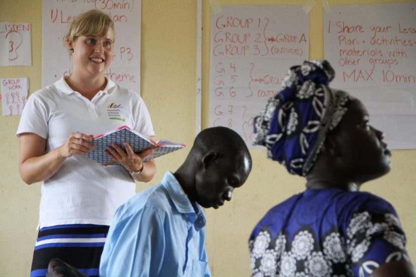 Seisova, hymyilevä vapaaehtoinen ja kaksi istuvaa ugandalaista katsoo kuvan ulkopuolelle. Seinällä on papereita, joissa on kirjoitusta.