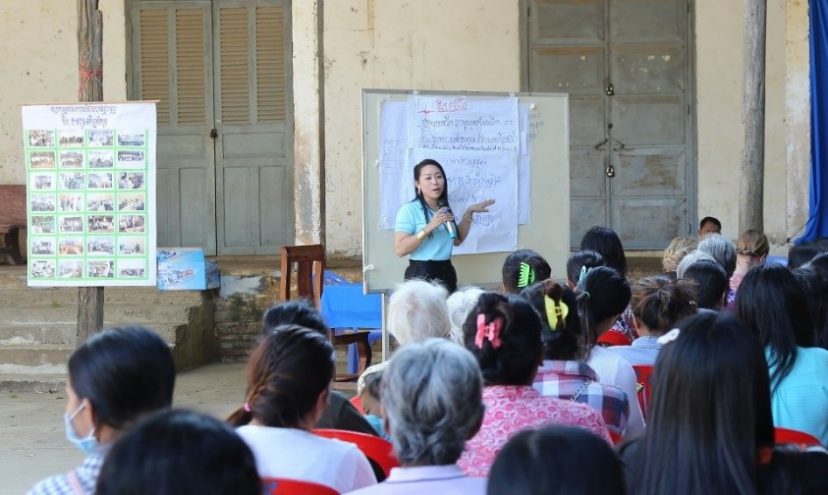 Kambodzalainen nainen puhuu isolle ihmisjoukolle ja osoittaa vieressä olevaa taulua, jossa on kirjoitusta.