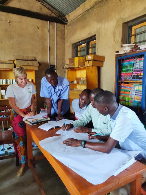 Vapaaehtoinen ja neljä ugandalaista opettajaa istuu tai seisoo pöydän ääressä, ja tarkastelee edessään olevaa paperia.