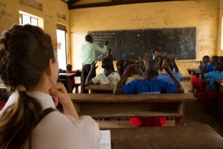 Vapaaehtoinen istuu ugandalaisen luokkahuoneen takaosassa ja seuraa opetusta.