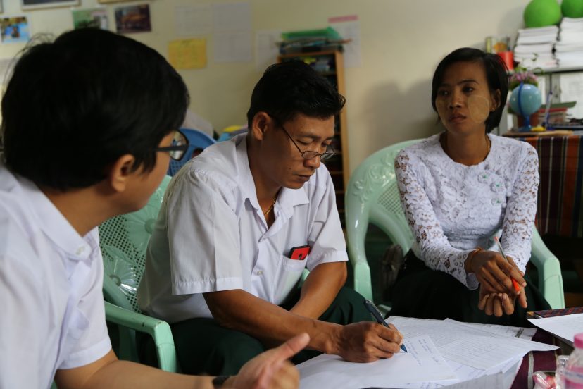 Kolme myanmarilaista henkilöä istuu ison paperin ääressä, johon yksi heistä kirjoittaa.