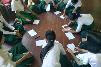 Students sit on floor in a ring with paper in front of them.