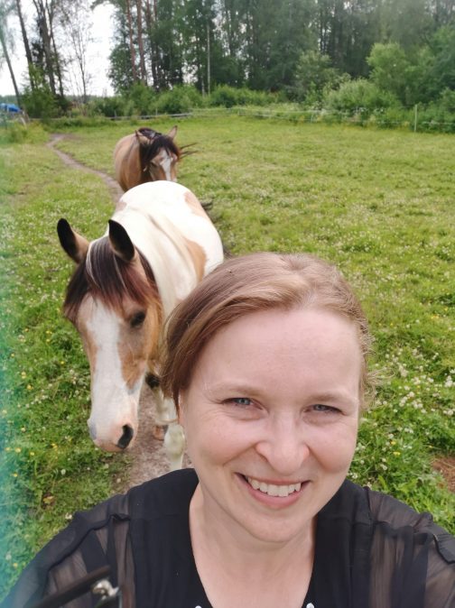 Nainen hymyilee kameralle takanaan kaksi hevosta.