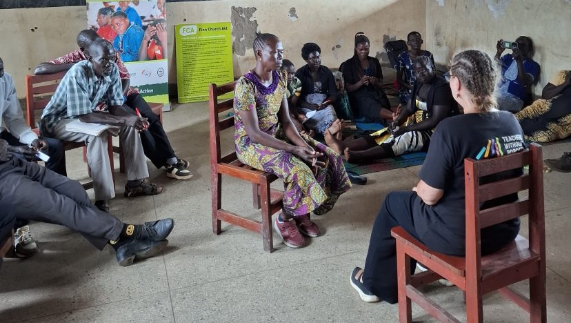 Teachers sitting in a circle, on the ground and chairs and discussing.