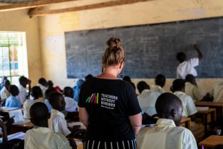 Luokkahuone, jossa oppilaat istuvat kohti taulua. Opettaja kirjoittaa taululle. Keskelläa takana seisoo nainen, jolla on musta t-paita, jossa lukee Teachers Without Borders